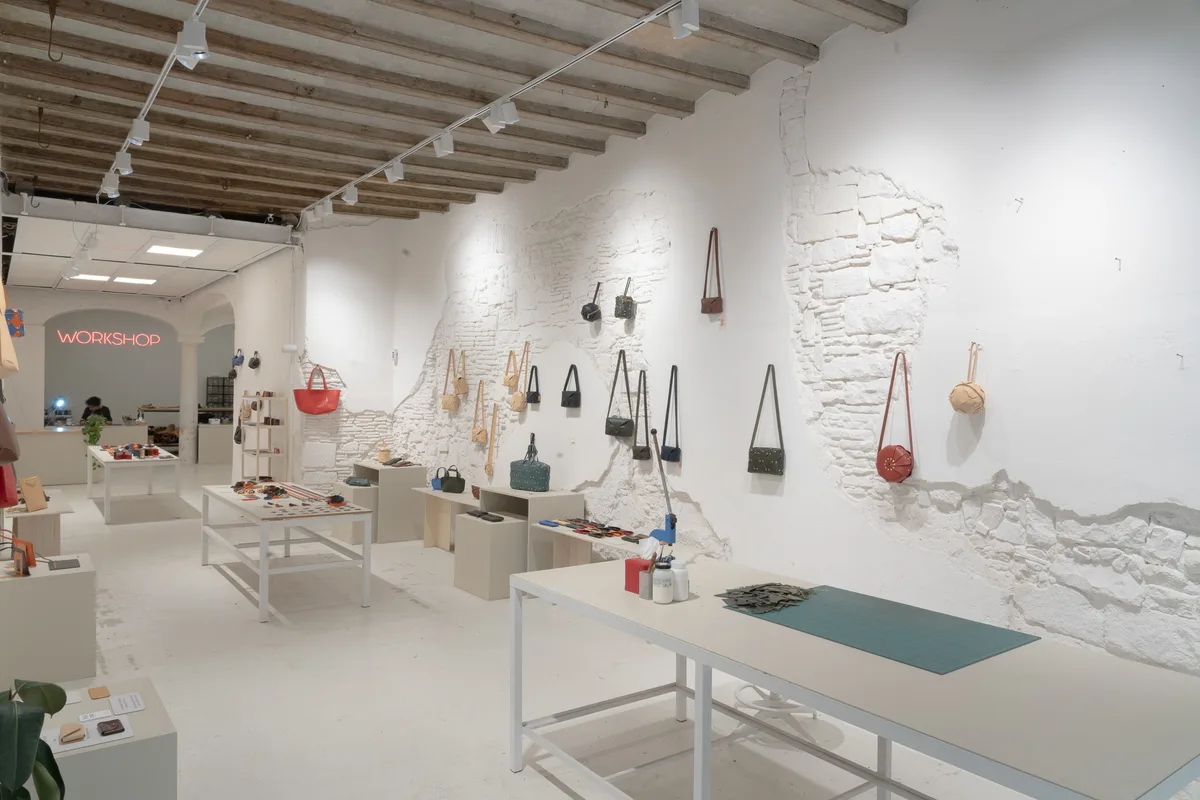 Interior of Atelier Madre with bags hung along the white stone wall and the WORKSHOP neon sign glowing at the far end