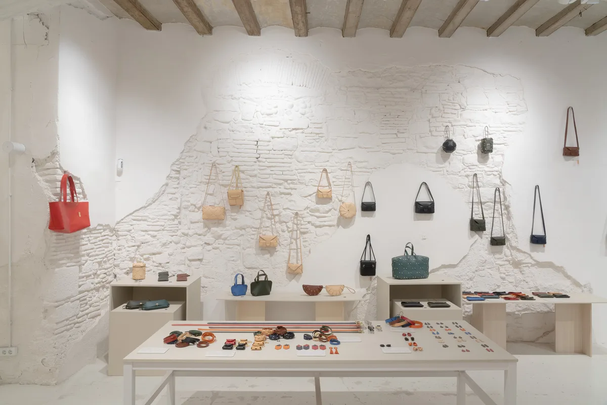 Taula central d'Atelier Madre amb arracades, polseres i carteres de pell, i bosses penjades del mur de pedra al fons