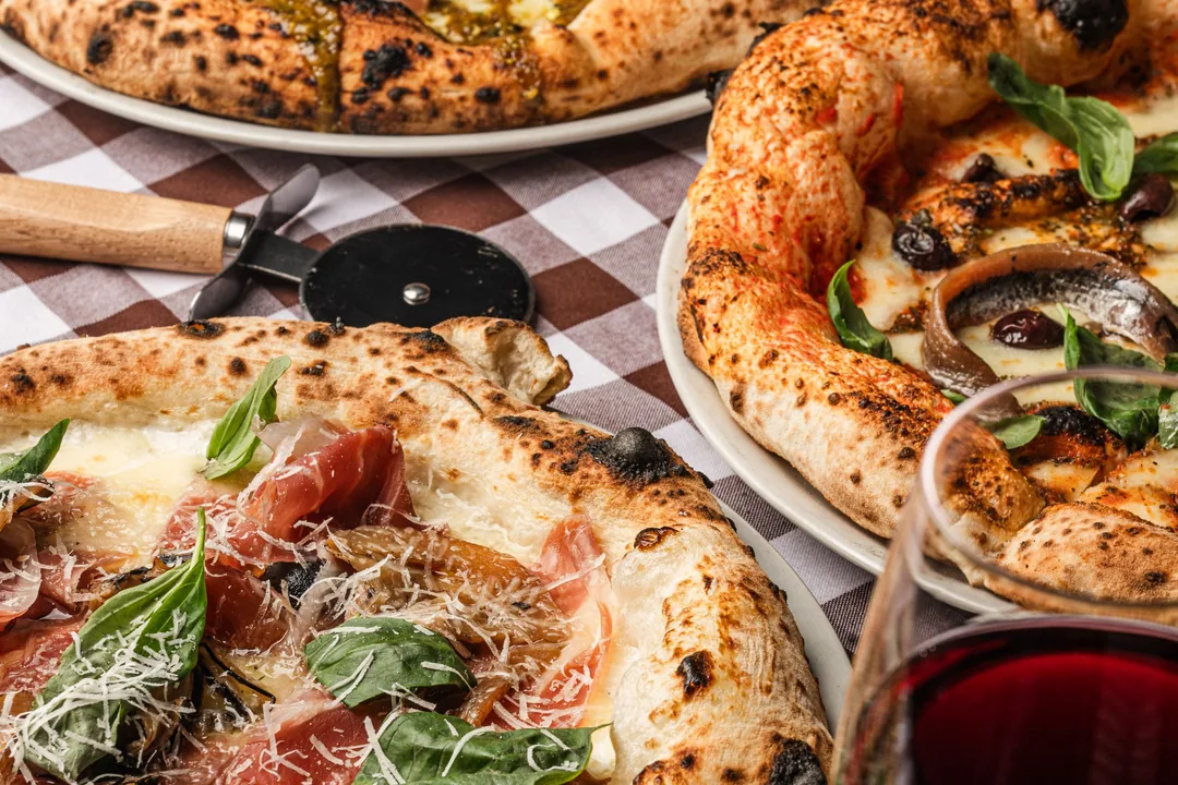Pizzas napolitanas con jamón y albahaca sobre un mantel de cuadros con una copa de vino tinto