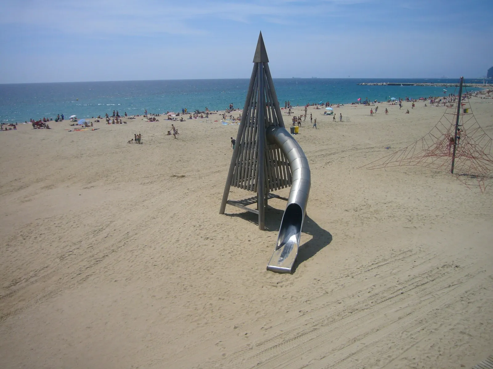 Arena de la platja de la Mar Bella con una escultura metálica en primer plano y el mar al fondo